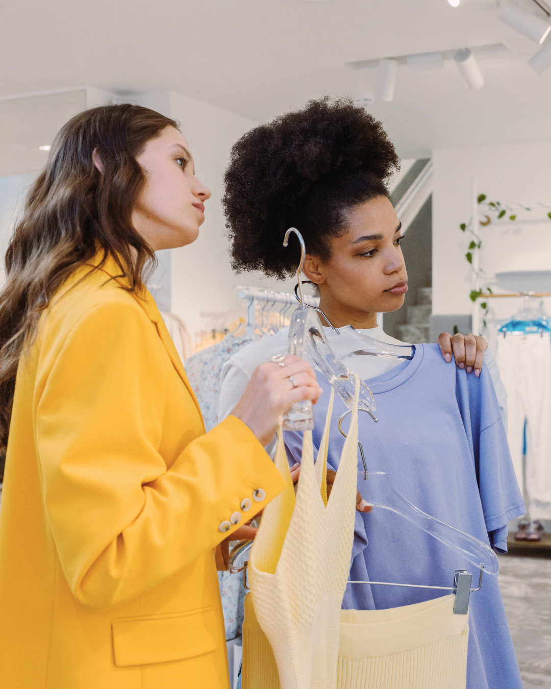 Two women comparing clothing pieces inside a fashion boutique while choosing an outfit.